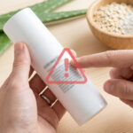 Une main tient un flacon cosmétique blanc avec un panneau d'avertissement rouge. Aloe vera et ingrédients naturels en fond.