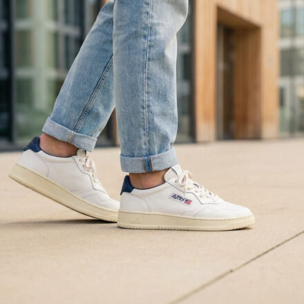 La basket Autry : l’icône vintage qui domine le style Gros plan sur une personne portant des baskets Autry blanches avec un empiècement bleu marine, des jeans clairs roulottés, marchant en ville.