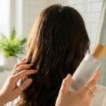 Vue de dos d'une personne aux cheveux bruns ondulés appliquant un produit capillaire depuis une bouteille en verre dans une salle de bain lumineuse.