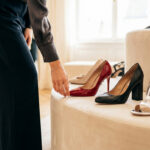 Vue rapprochée d'une personne en pantalon sombre touchant un talon aiguille rouge laqué, entourée d'autres chaussures élégantes en boutique.