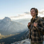 Jeune personne souriante avec un sac à dos, admirant un vaste paysage montagneux et une vallée profonde sous un ciel lumineux.
