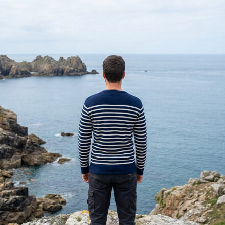 Armor-Lux : le savoir-faire breton d’un vestiaire durable Un homme en marinière rayée contemple la mer et les falaises rocheuses de Bretagne. Un phare se dresse au loin sur une pointe.