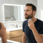 Homme barbu taillant précisément sa barbe avec une tondeuse électrique devant un miroir éclairé dans une salle de bain moderne.