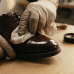 Une personne gantée polit le bout brillant d'une chaussure en cuir marron avec un chiffon. Pot de cirage et brosse sur table en bois.