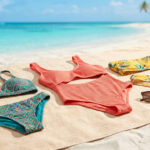 Trois maillots de bain (bikini turquoise, une pièce corail, bikini floral jaune) et lunettes sur serviette de plage tropicale.
