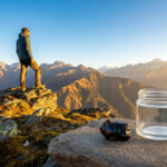 Un morceau de shilajit et un pot vide sur un rocher, avec un randonneur admirant une chaîne de montagnes au lever du soleil.
